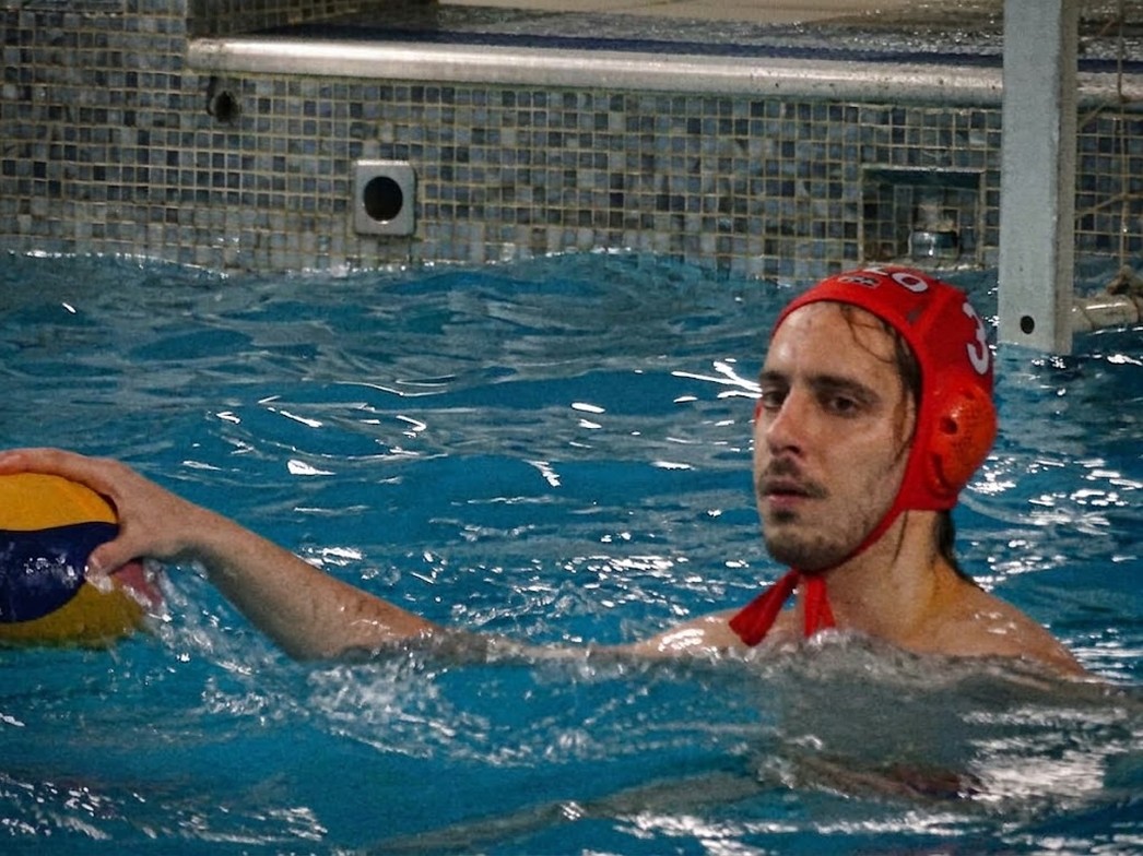 Giacomo playing water polo in a pool, wearing a red cap